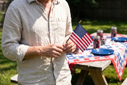 Man holding small American flag at a backyard Fourth of July picnic with patriotic table setting, plates, and summer decorations