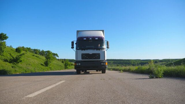Front view of truck with cargo trailer driving on highway transporting goods at summer day. White lorry riding through countryside with beautiful nature landscape at background. Slow motion Front view