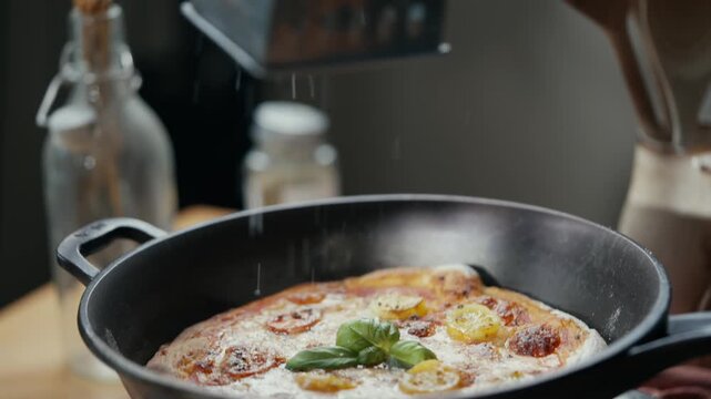 Hand grates cheese over pizza in skillet. Warm kitchen scene with rustic decor. Slow motion captures cheese falling. Ideal for food, cooking, or lifestyle videos. Focus on culinary action and texture