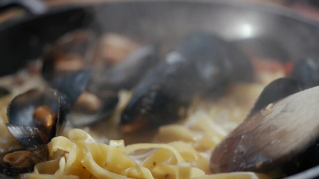 Steaming pasta with mussels in a pan. Wooden spoon stirs savory dish. Close-up shot captures delicious food details. Ideal for cooking, restaurant, or food-related projects