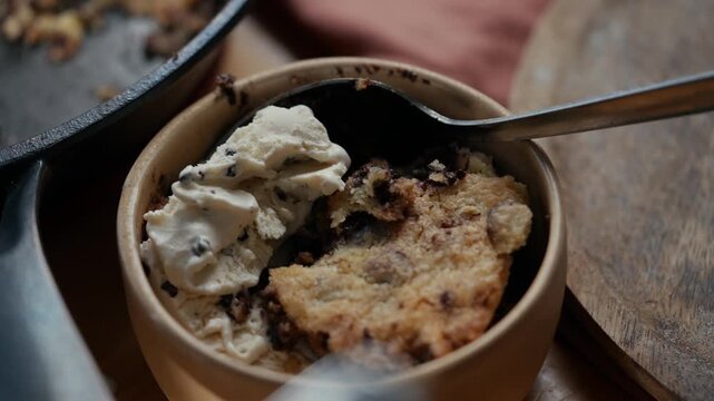 Warm cookie dough dessert with creamy vanilla ice cream in rustic ceramic bowl. Scoop rests invitingly in soft, crumbly treat. Background shows cast-iron skillet with scattered crumbs