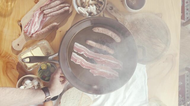 Top-down view shows hands placing bacon and sausages into a steaming skillet. Ingredients like mushrooms, butter, and greens surround the pan. Steam rises as meat sizzles