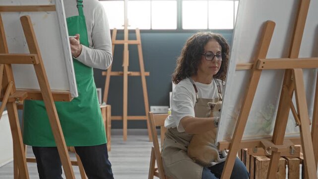 Man holding paintbrush paints on canvas in sunlit art studio, standing beside seated woman artist in apron; creative companionship.