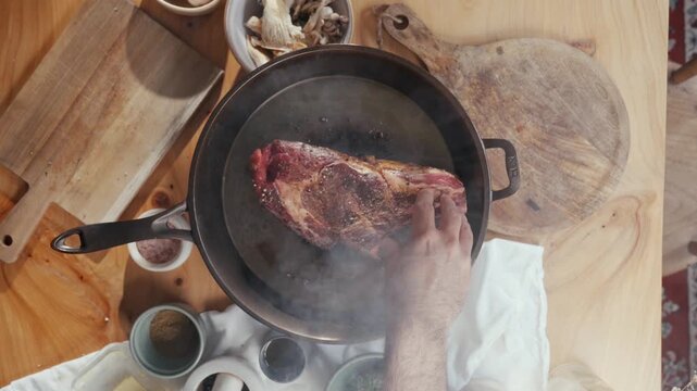 Hand places raw steak into hot pan. Steam rises as meat hits oil. Wooden table holds spices and tools. Scene shows careful cooking preparation. Ideal for food, restaurant, or cooking tutorials