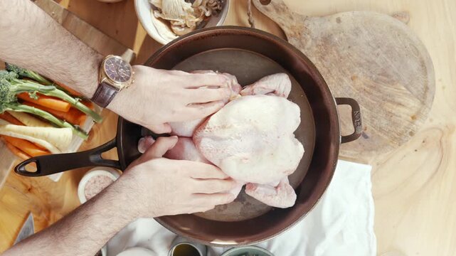 Hands place raw chicken in pot. Ingredients like carrots and herbs surround pot. Scene shows meal prep in rustic kitchen. Ideal for cooking tutorials or food blogs. Emotion: focused, calm, methodical
