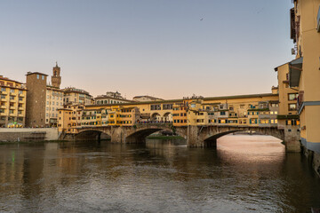 Fototapeta premium The iconic Ponte Vecchio bridge in Florence, Italy, glows with warm light during a serene sunset over the Arno River