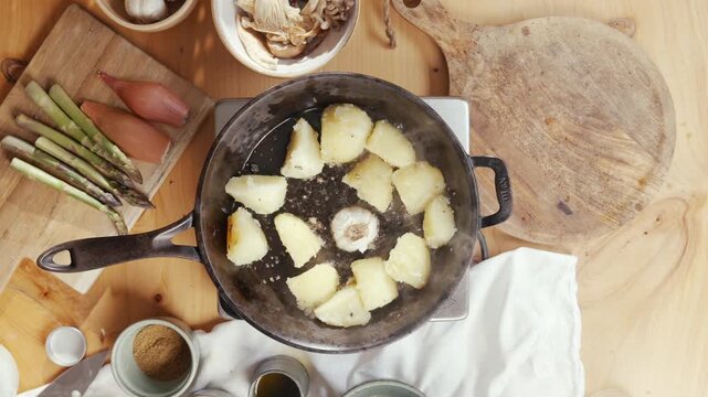 Golden potato chunks fry in a cast-iron skillet. Wooden spatula stirs them gently. Steam rises as they brown in hot oil. Kitchen counter holds fresh ingredients nearby