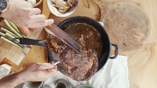 Hand lifts juicy, roasted meat from a pot. Rich brown gravy and fresh ingredients like garlic, shallots, and mushrooms. The person is focused, carefully handling the meat with care