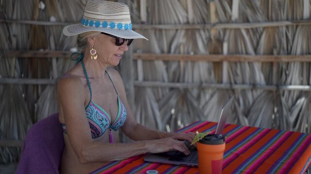 Mature woman works on computer with phone nearby on tropical beach while displaying different emotions during her vacation time.