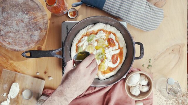 Hand pours olive oil over a fresh pizza in a cast-iron pan. Ingredients like mozzarella and tomato sauce are visible. The scene is warm and inviting, showing home cooking with care