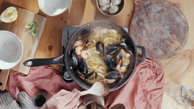 Hand stirs mussels in a skillet over a wooden table. Warm kitchen light highlights fresh ingredients and rustic cloth. Cooking scene evokes comfort and home-style meal preparation