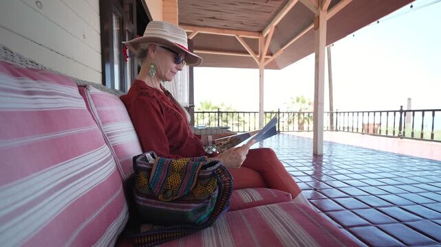 A mature woman sits on a veranda in Santa Rosalia Baja California Sur and looks at a magazine and a guidebook during her travel.
