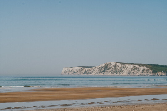Sunlit beach meets calm sea under clear blue sky. White cliffs stand tall, rugged and serene. Sand stretches wide, soft and inviting. Waves gently roll in, creating quiet rhythm