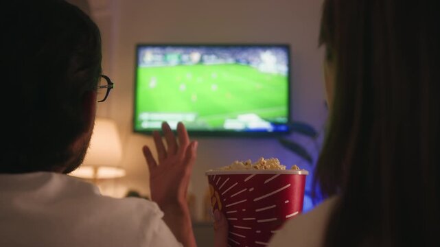 Couple watching a football match on TV at home. Disappointed fan throws popcorn toward the screen after a missed scoring chance. Sports emotions, frustration and football fandom concept. High quality
