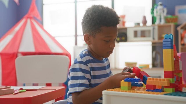 Boy stacking colorful plastic blocks with hands at a low table in a bright playroom inside a building, toy tent and shelves nearby; creative concentration.