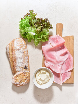 Sandwich preparation with ciabatta bread, ham, lettuce, and mayonnaise viewed from above on a neutral background