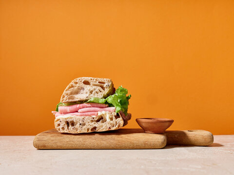 Ciabatta sandwich with ham and lettuce on a wooden board against an orange background featuring bright colors and copy space