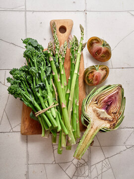 Fresh kale, broccolini, artichoke, asparagus, and kumato tomatoes arranged on a wooden board in a kitchen setting