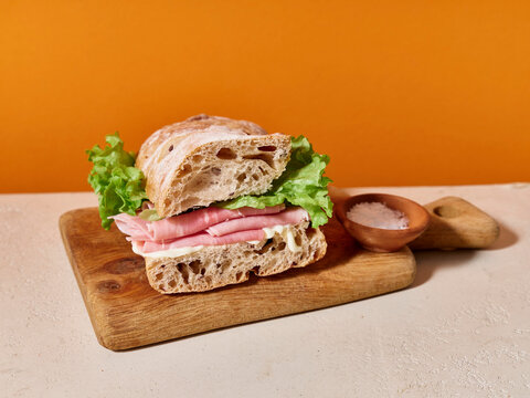 Ciabatta sandwich with ham, lettuce, and mayonnaise on a wooden board against a bright orange background