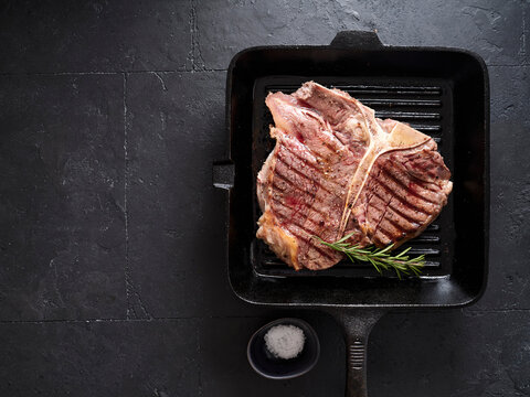 Grilled t-bone steak on a cast iron skillet with a sprig of rosemary and a small bowl of salt on a dark surface