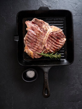 Grilled t-bone steak on a grill pan with salt and rosemary on a dark stone background