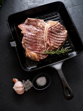 Grilled t-bone steak cooking in a skillet with garlic and seasoning on a dark kitchen surface