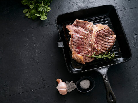 Grilled t-bone steak on a cast iron skillet with herbs and garlic on a dark stone surface