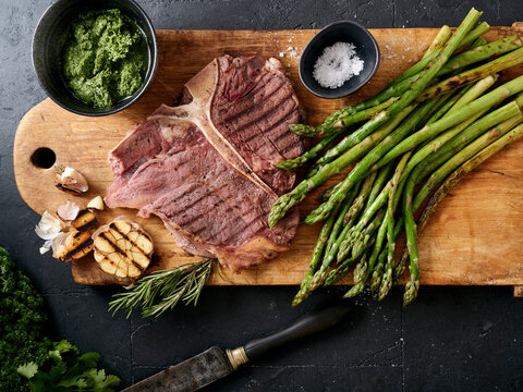 Grilled t-bone steak with asparagus and garlic on a wooden cutting board with sauces and herbs arranged around the meat