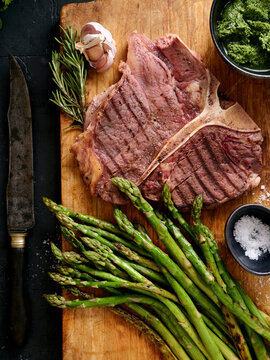 Grilled t-bone steak with fresh asparagus and herbs on a wooden cutting board in a kitchen setting during the day