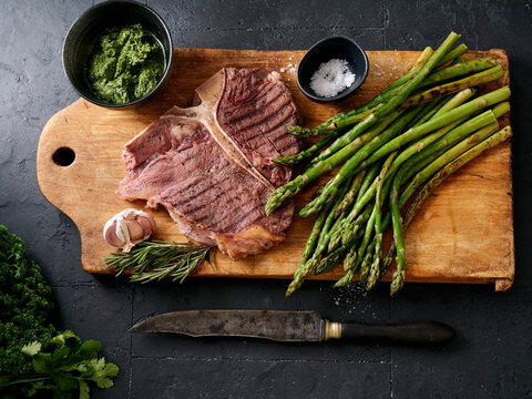 Grilled t-bone steak with asparagus and sauces prepared on a wooden board with garlic and herbs in a kitchen setting