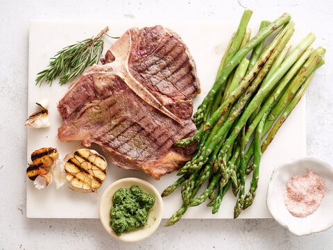 Grilled t-bone steak served with asparagus and garlic on a cutting board near fresh herbs and sauce