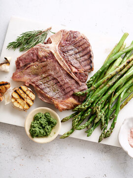 Grilled t-bone steak served with asparagus and garlic on a wooden board in a kitchen setting during daylight