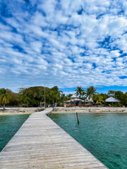Fototapeta premium Wooden pier leading to tropical beachfront villas with palm trees and turquoise Caribbean water under dramatic cloudy sky in Utila, Honduras.