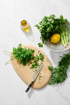 Overhead shot of fresh parsley (partly chopped), coriander, dill, lemon and olive oil on a white marble surface with a knife, board and colander, showing ingredients preparation