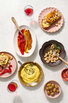 Angled shot of a bowl of braised butter beans with rosemary and lemon peel, some beans served onto toasted sourdough with saffron aioli and charred peppers, with plates of saffron aioli.