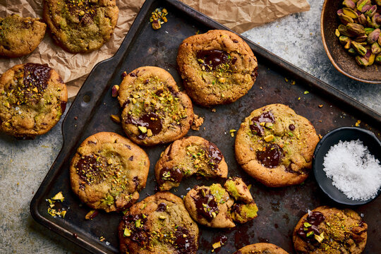 Pistachio-studded chocolate cookies with molten centers arranged on a dark, weathered baking tray, with scattered nuts, flaky salt, parchment paper, and a ceramic bowl of pistachios.