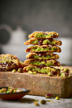 A vertical stack of pistachio-stuffed chocolate cookies split open to reveal a vibrant green crumb and molten chocolate, styled on a rustic wooden cutting board with scattered pistachios and crumbs.