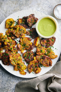 Crispy, golden smashed potatoes roasted with grated parmesan cheese and fresh herbs, served alongside a vibrant green dipping sauce with lime. Styled on a rustic custom stone surface.