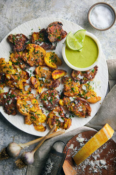 Crispy, golden smashed potatoes roasted with grated parmesan cheese and fresh herbs, served alongside a vibrant green dipping sauce with lime. Styled on a rustic custom stone surface.