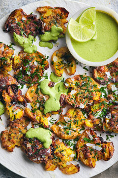 Close up of crispy, golden smashed potatoes roasted with grated parmesan cheese and fresh herbs, drizzled with a vibrant green dipping sauce with lime.