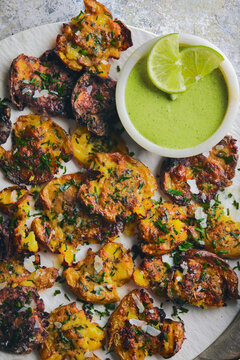 Close up of crispy, golden smashed potatoes roasted with grated parmesan cheese and fresh herbs, served alongside a vibrant green dipping sauce with lime.