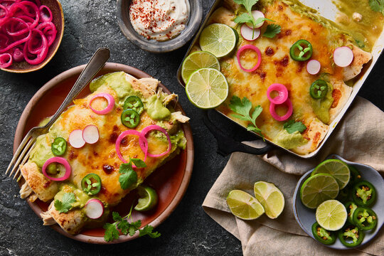 Golden-baked enchiladas layered with melted cheese and green sauce, topped with fresh cilantro, jalape&ntilde;os, radishes, and pickled red onions, served with lime wedges.