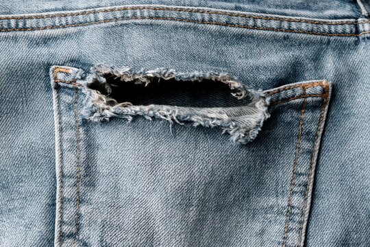 Close-up detail of a ripped pocket on worn blue denim jeans, showing frayed threads and texture.
