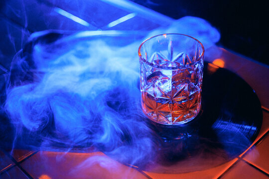 Blue smoke whiskey glass on vinyl record with dark neon nightlife mood. Concept of smoke drink hero shot, premium whiskey campaign, music bar poster, speakeasy mood, and social cover.