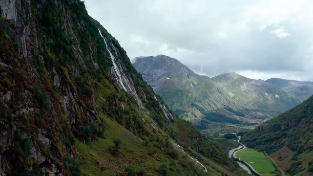 High-angle aerial drone footage reveals a majestic waterfall cascading into a mountain river valley in Klakegg, Norway. Beautiful summer landscape shows cars driving on a highway in Summer