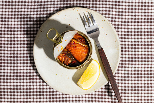 Open tin of salmon with lemon and fork on plate in hard light