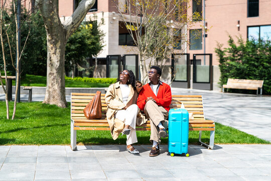 Cheerful curious african american couple man and woman travelers resting together on park bench in new city, smiling black male and female enjoying sunny weather while having break before flight.