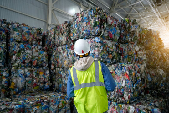 Worker in reflective vest and white helmet stands against a background of stacks of compressed pressed tetra packs cardboard, folded into cubes packaging briquettes waste processing plant warehouse