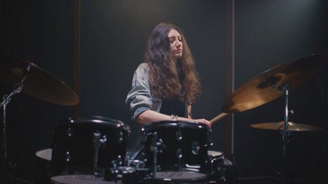 Young drummer girl counts down rhythm with drumsticks, and song starts. Female musician with long curly hair plays a drum kit. Rock band on concert, live on rehearsal room. Repetition in slow motion