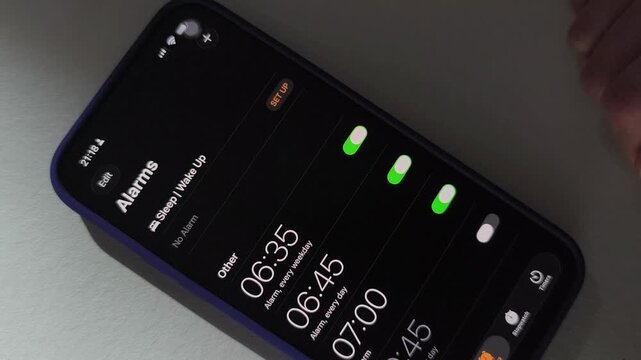 Hand taps smartphone alarm settings on dark screen. Finger enables wake up toggles and adjusts morning schedule. Mobile time management and routine planning concept..